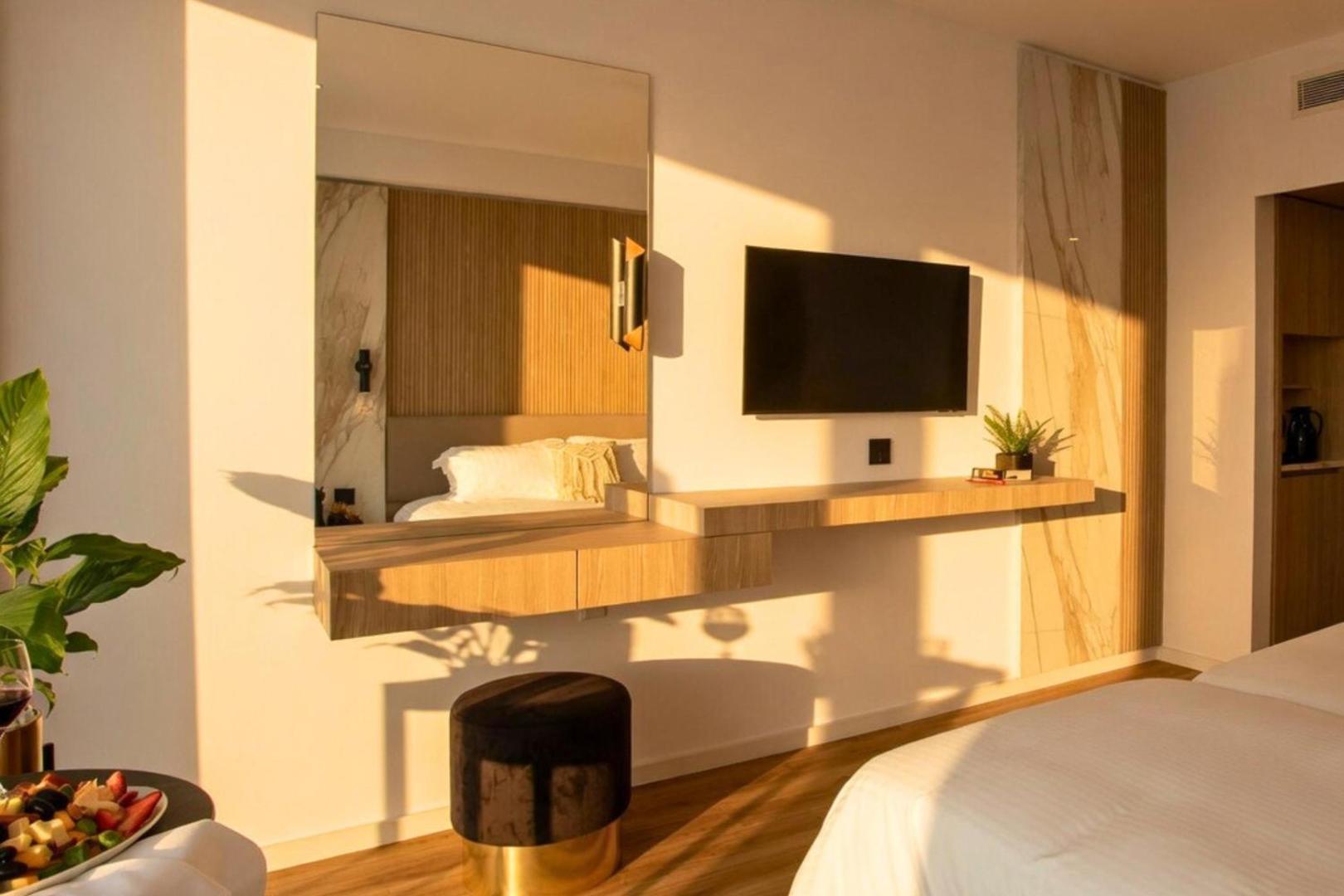 Cavo Maris Beach numura interjera detaļas — darba zona ar spoguli, TV un koka apdari siltā pēcpusdienas saulē
