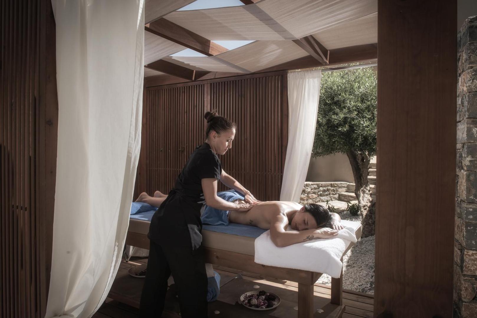 Spa procedūra Enorme Infinity Elounda viesnīcas āra spa zonā, masāža koka konstrukcijas lapenē ar baltiem aizkariem