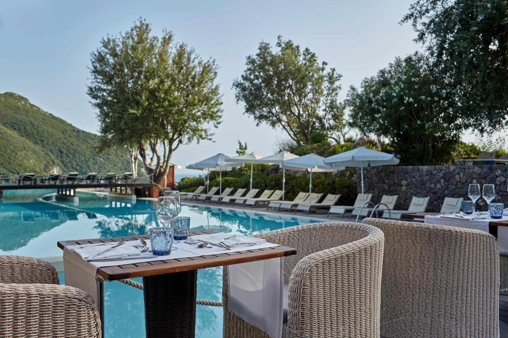 Atlantica Grand Mediterraneo Resort āra baseins ar sauļošanās krēsliem, saulessargiem un galdiņu priekšplānā Korfu piekrastē