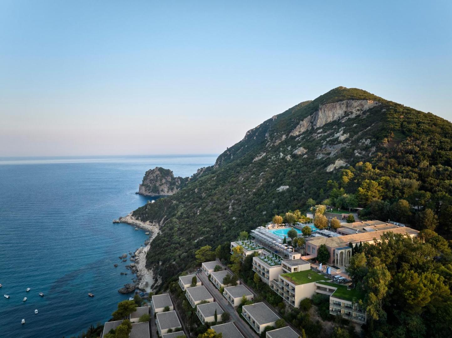Atlantica Grand Mediterraneo Resort skats no augšas uz galveno ēku, āra baseinu un bungalo tipa numuriem kalna nogāzē saulrietā.
