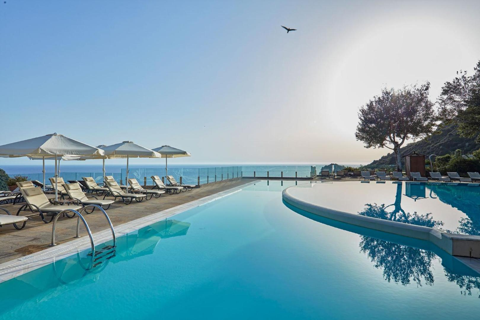Atlantica Grand Mediterraneo Resort āra baseina zona ar sauļošanās krēsliem un saulessargiem pie stikla margām ar skatu uz jūru.