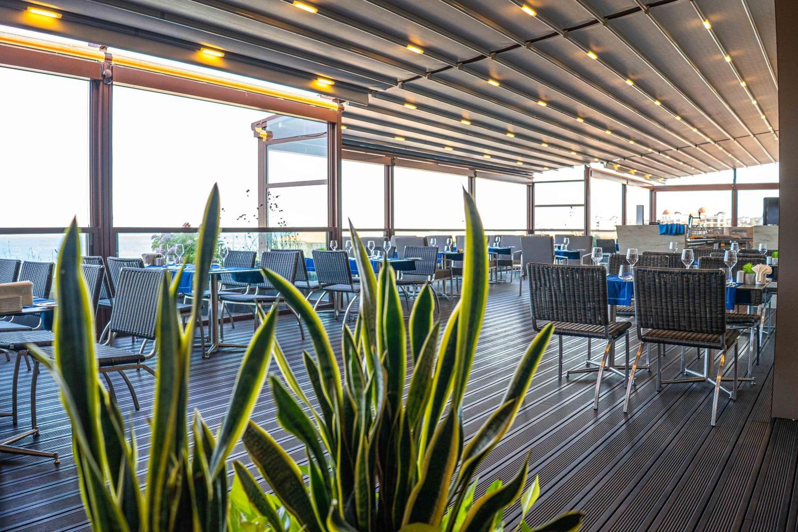 Skats uz HVD Club Hotel Miramar restorāna terasi caur telpaugiem — modernas mēbeles un bīdāmā jumta konstrukcija komfortablai atpūtai.