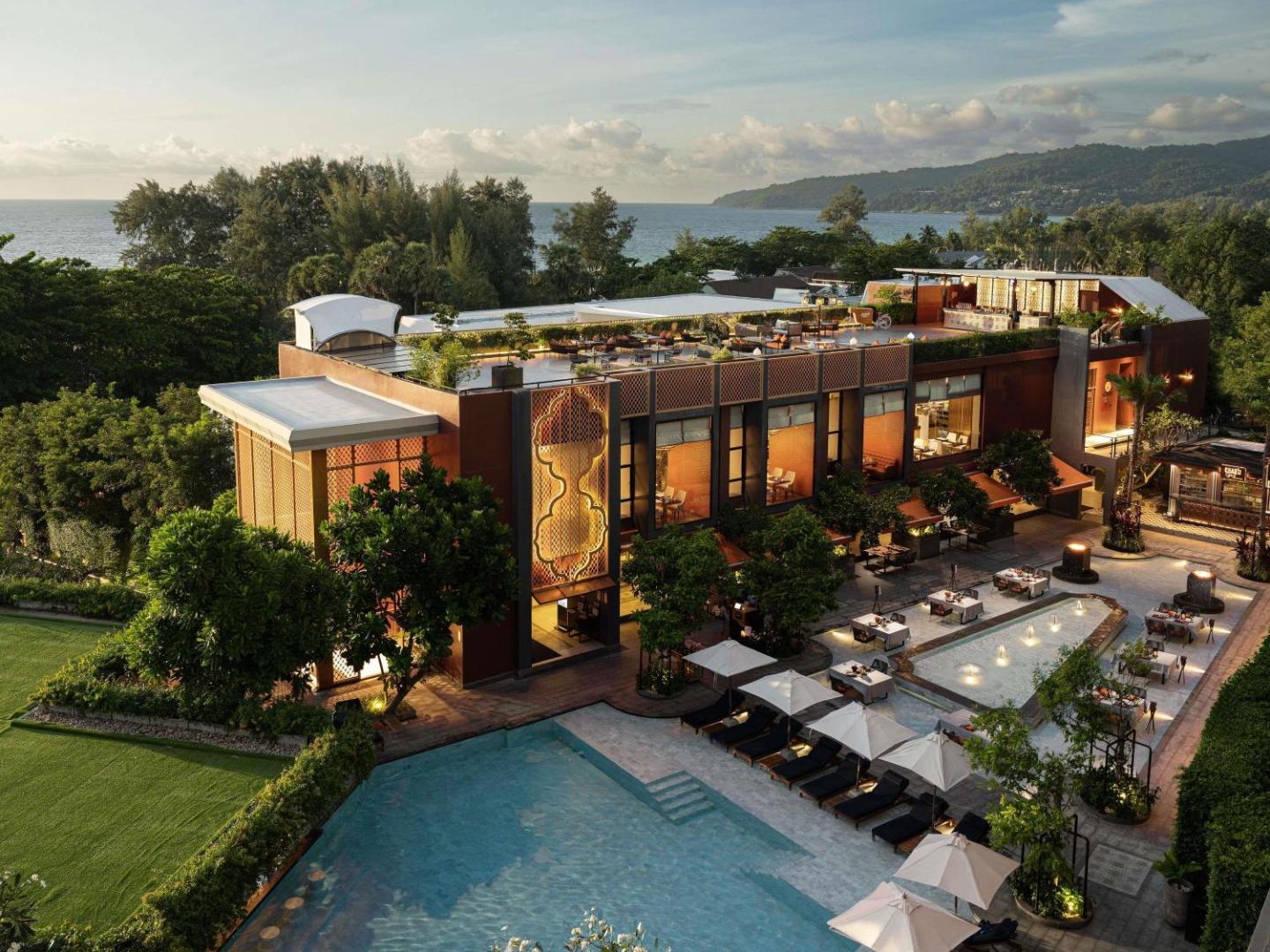 Avista Grande Phuket skats no augšas uz āra baseinu, restorāna terasi un viesnīcas ēku saulrietā Karon piekrastē, Taizemē.