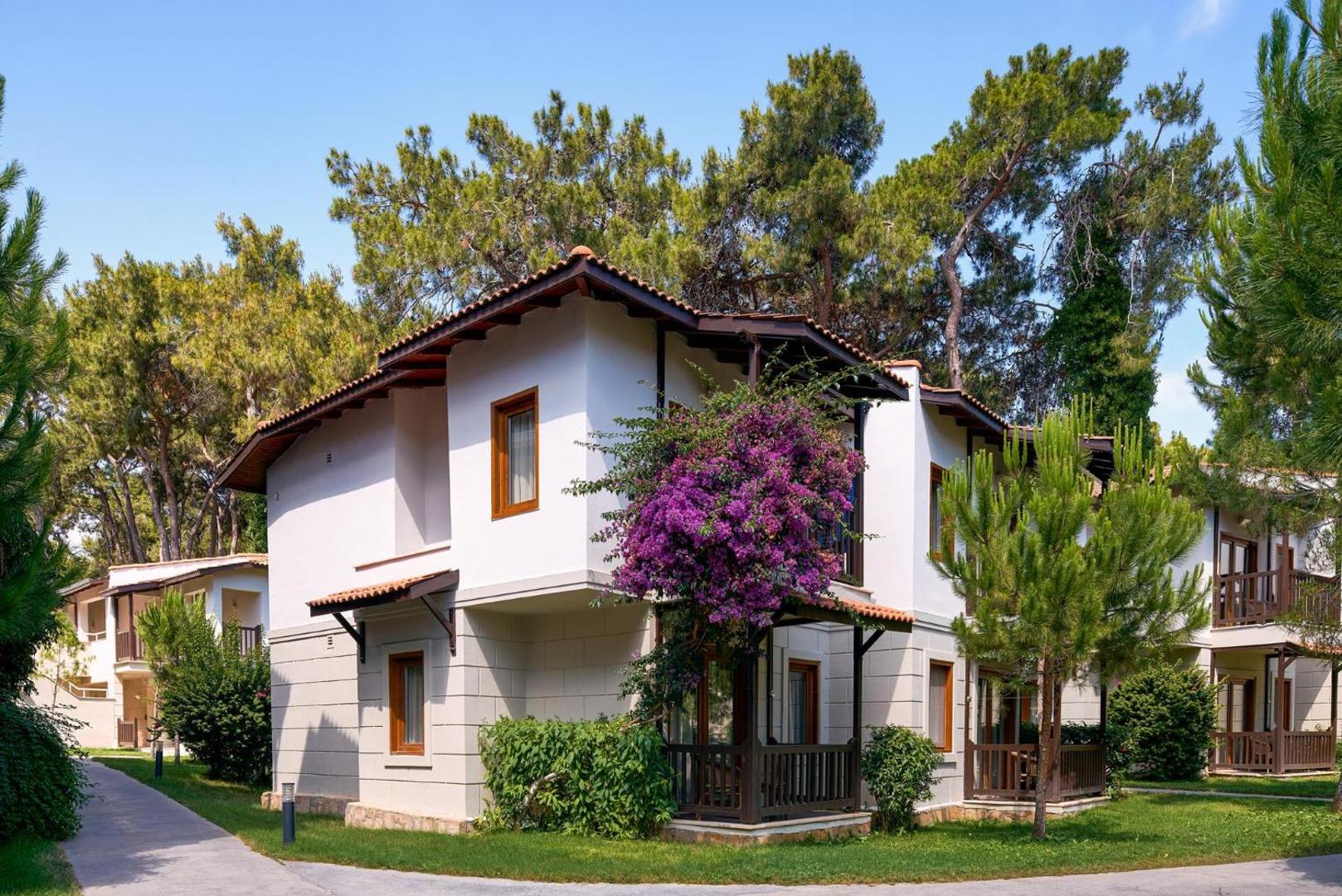 Voyage Sorgun divstāvu bungalo tipa numuri ar balkoniem, ko ieskauj ziedoši krūmi un augstas priedes