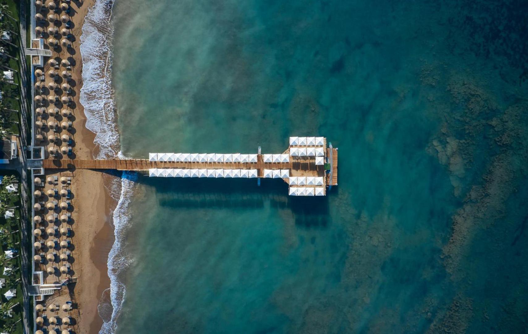 Aerofoto skats uz Voyage Sorgun viesnīcas koka laipu un smilšu pludmali ar saulessargiem dzidri zilā Vidusjūras piekrastē
