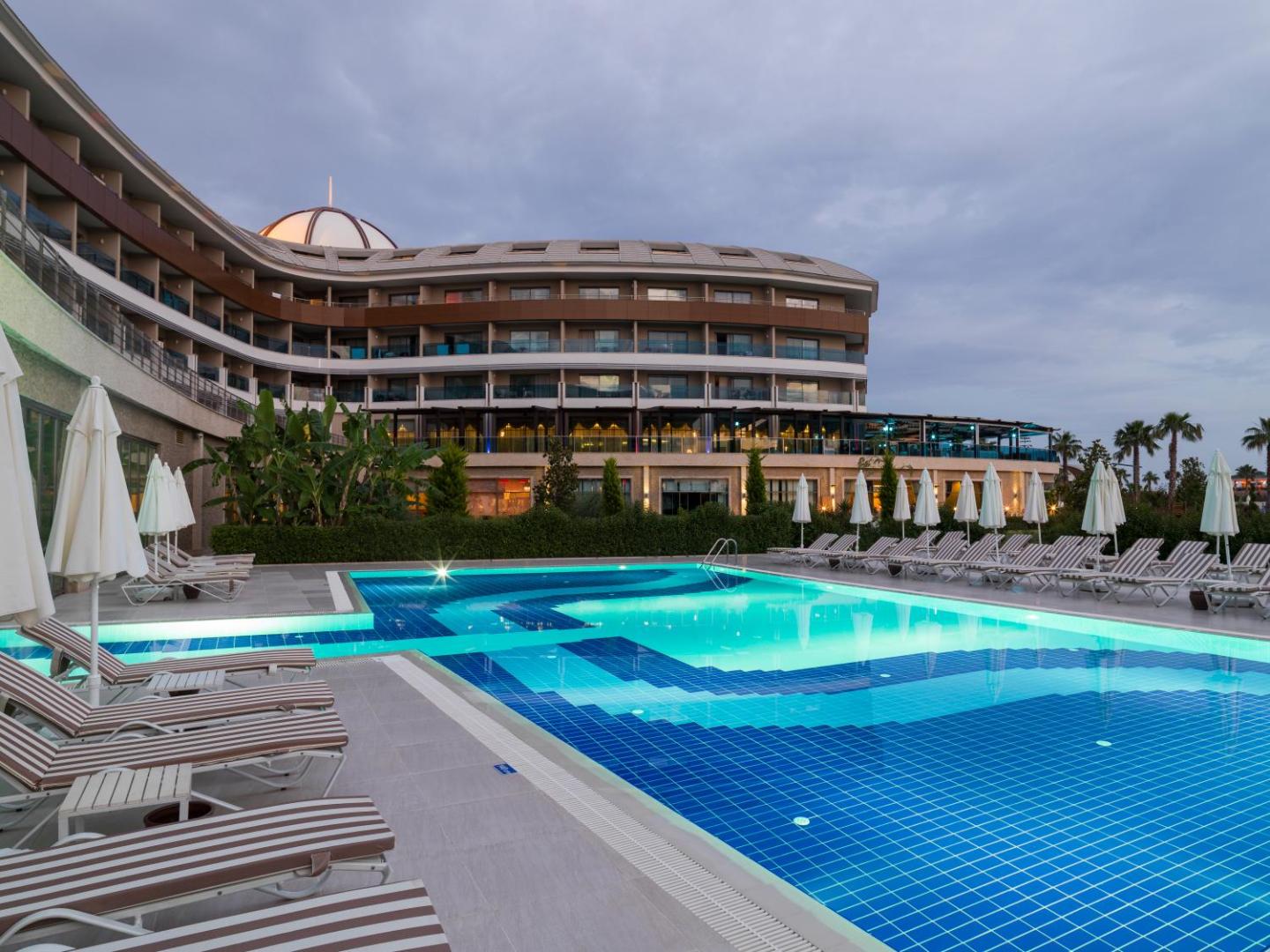 TUI Magic Life Jacaranda āra baseins vakarā ar apgaismojumu un sauļošanās krēsliem — skats uz viesnīcas galveno ēku krēslas stundā