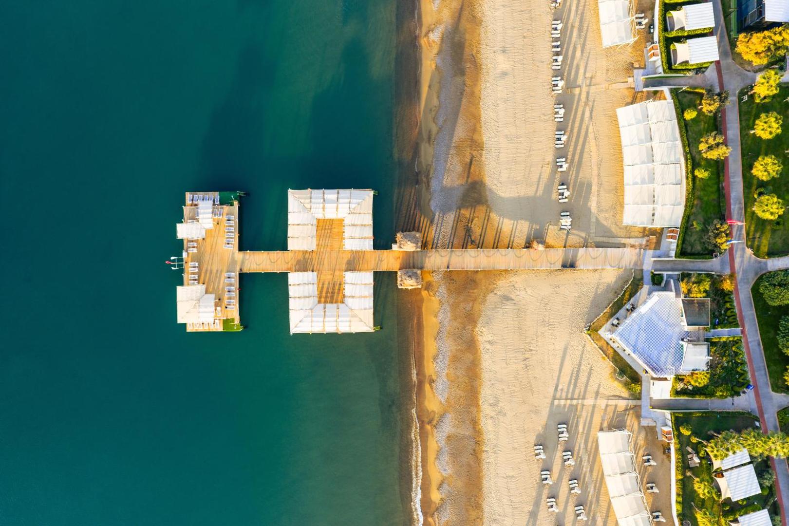 Susesi Luxury Resort koka piestātne ar atpūtas nojumēm un sauļošanās krēsliem virs tirkīzzilā jūras ūdens Belekā