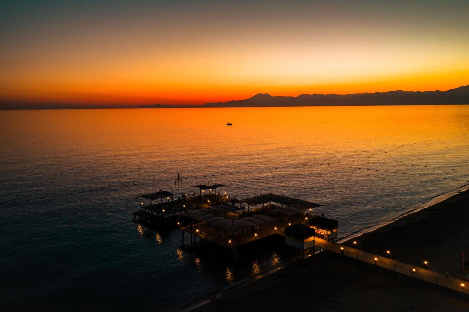 Susesi Luxury Resort privātā piestātne un bārs jūrā saulrieta laikā, oranžas debesis un kalnu silueti tālumā