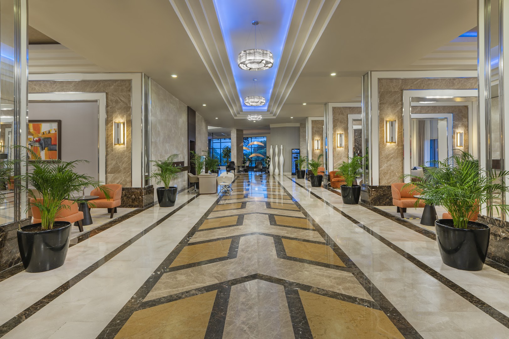 Susesi Luxury Resort plašais lobby gaitenis ar marmora rakstiem, oranžiem atpūtas krēsliem, telpaugiem un modernu apgaismojumu