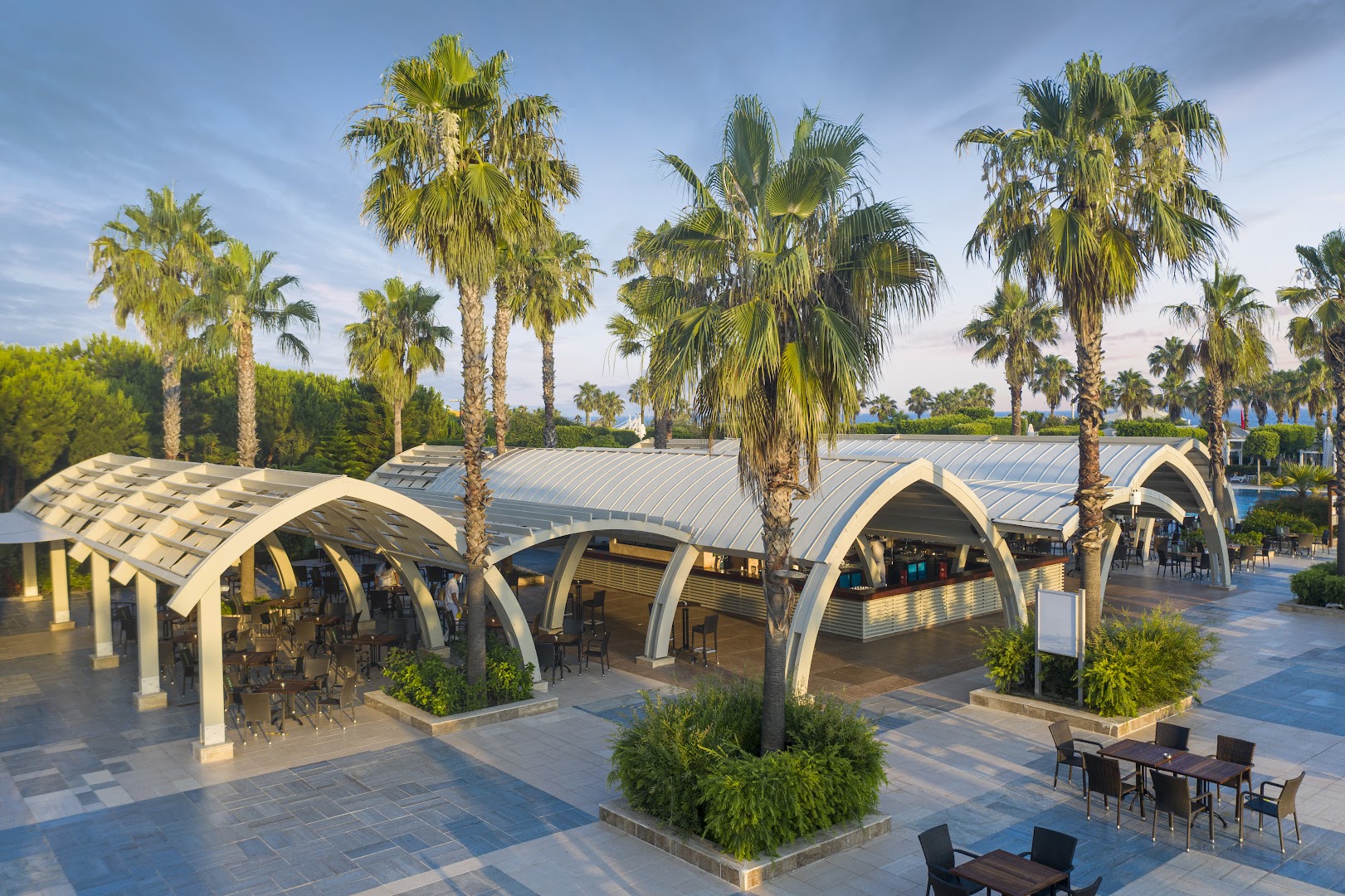 Susesi Luxury Resort āra bārs un atpūtas zona Belekā ar modernām arkveida nojumēm, palmām un galdiņiem saulainā pēcpusdienā