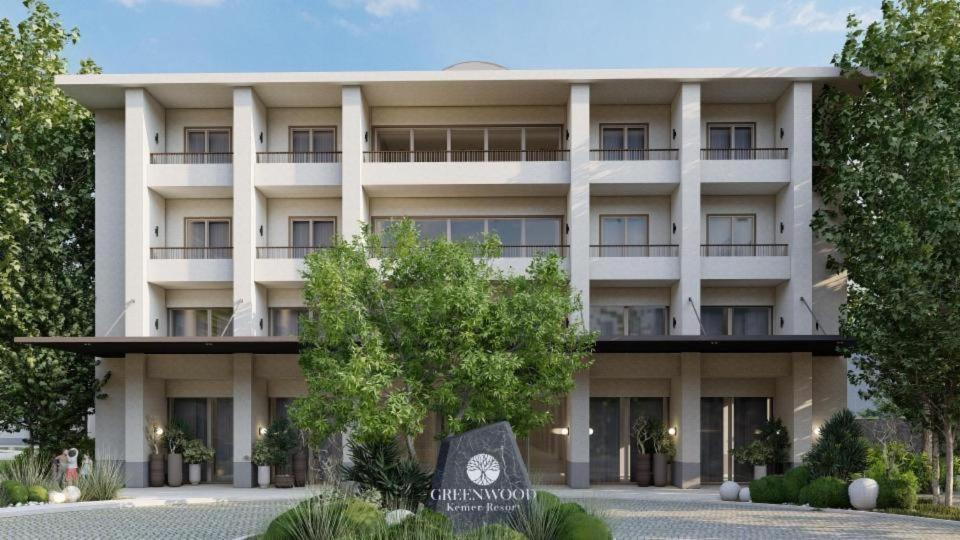 Greenwood Kemer Resort modernā korpusa fasāde ar balkoniem un ieeju, ko ieskauj zaļi koki un dekoratīvi augi.