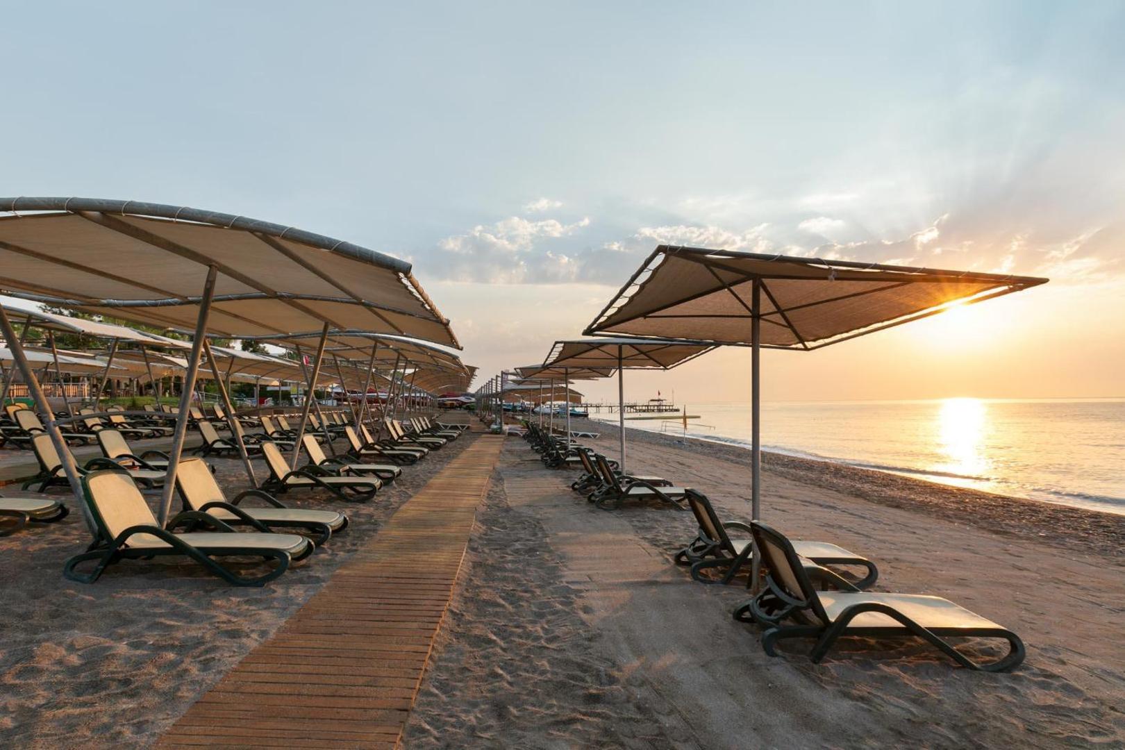Greenwood Kemer Resort privātā pludmale saulrieta laikā ar rindiņām sauļošanās krēslu zem nojumēm pie Vidusjūras