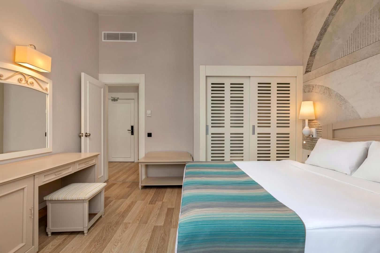 Greenwood Kemer Resort elegants numura interjers ar gaišām mēbelēm, dekoratīvu sienas krāsojumu un koka grīdu.