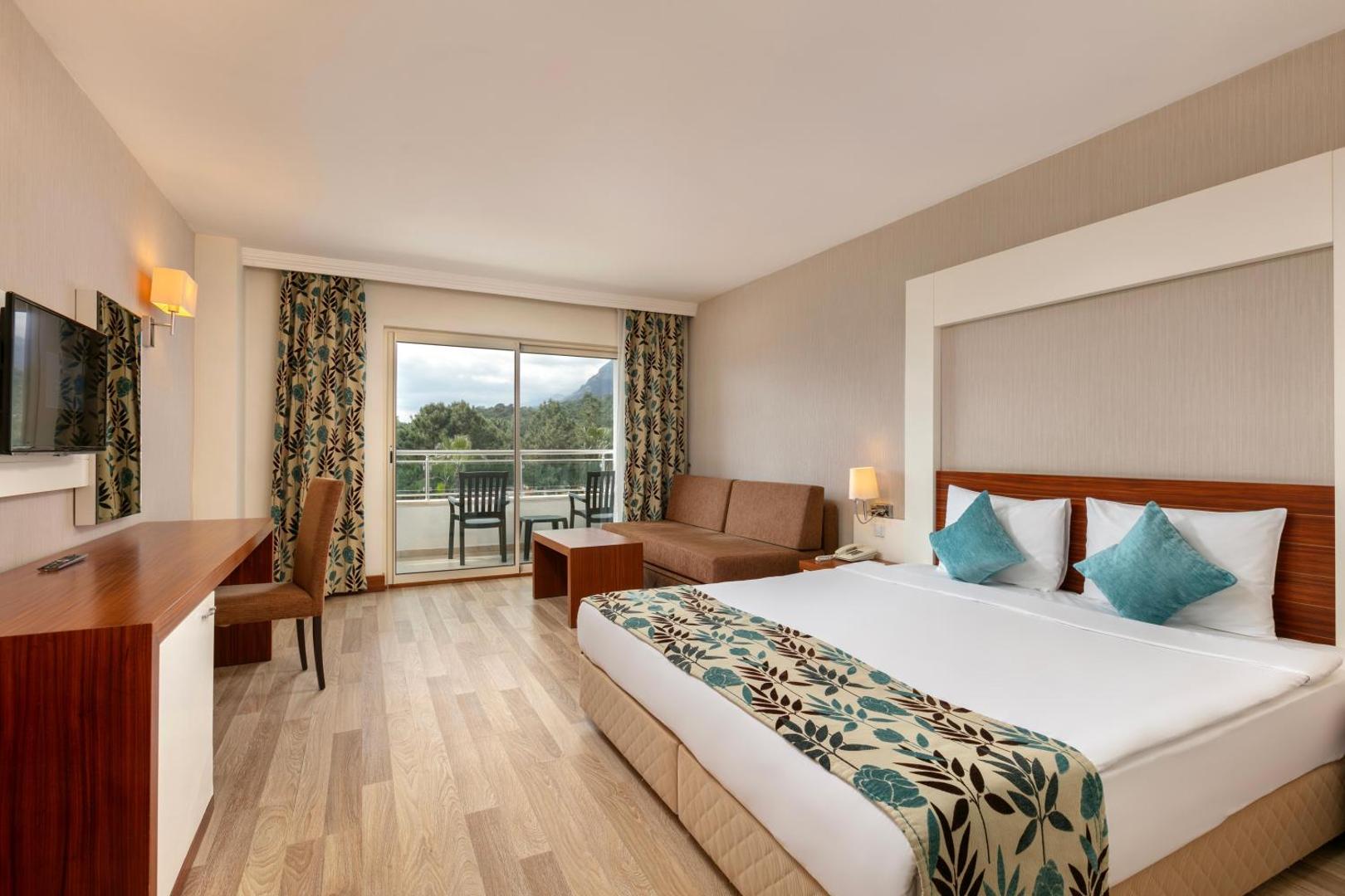 Gaišs Greenwood Kemer Resort viesnīcas numurs ar divguļamo gultu, dīvānu un balkonu, no kura paveras skats uz kalniem.