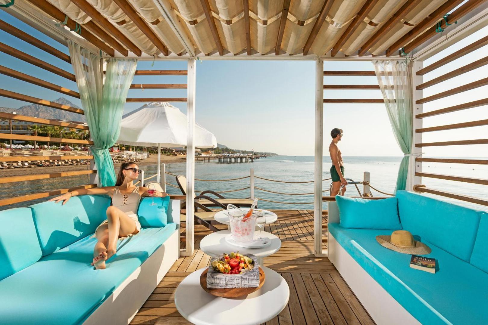 Ekskluzīva atpūtas nojume uz Sherwood Exclusive Kemer piestātnes ar mīkstiem dīvāniem, augļiem un tiešu skatu uz jūru un kalniem.