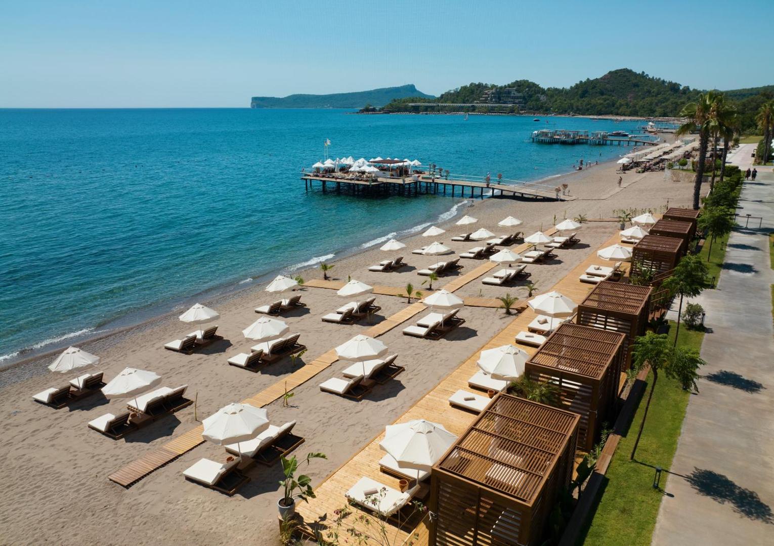 Sherwood Exclusive Kemer privātā pludmale no putna lidojuma ar saulessargiem, sauļošanās krēsliem, koka laipām un garu piestātni jūrā.