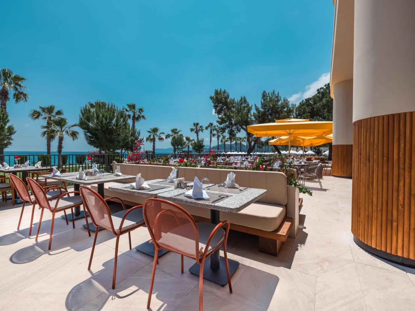 Sherwood Exclusive Kemer āra terases restorāns ar skatu uz jūru, klātiem galdiem, oranžiem krēsliem un dzelteniem saulessargiem palmām ieskautā vidē.