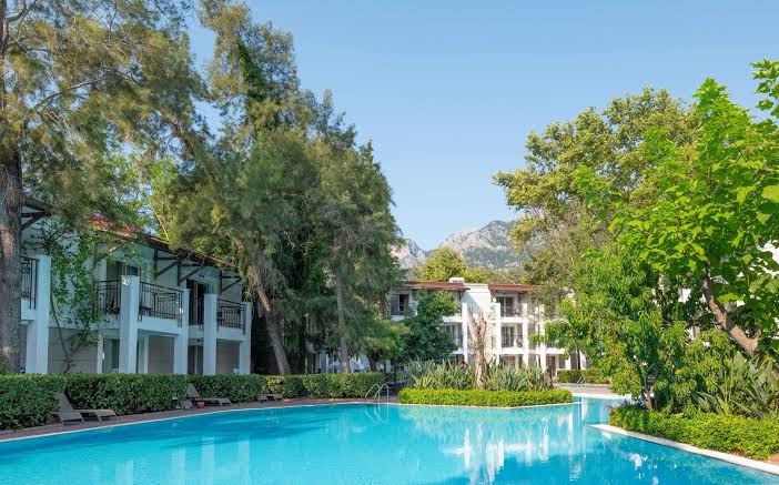Sherwood Exclusive Kemer viesnīcas korpusi ar balkoniem pie liela, dzidra āra baseina, ko ieskauj zaļi koki un kalni