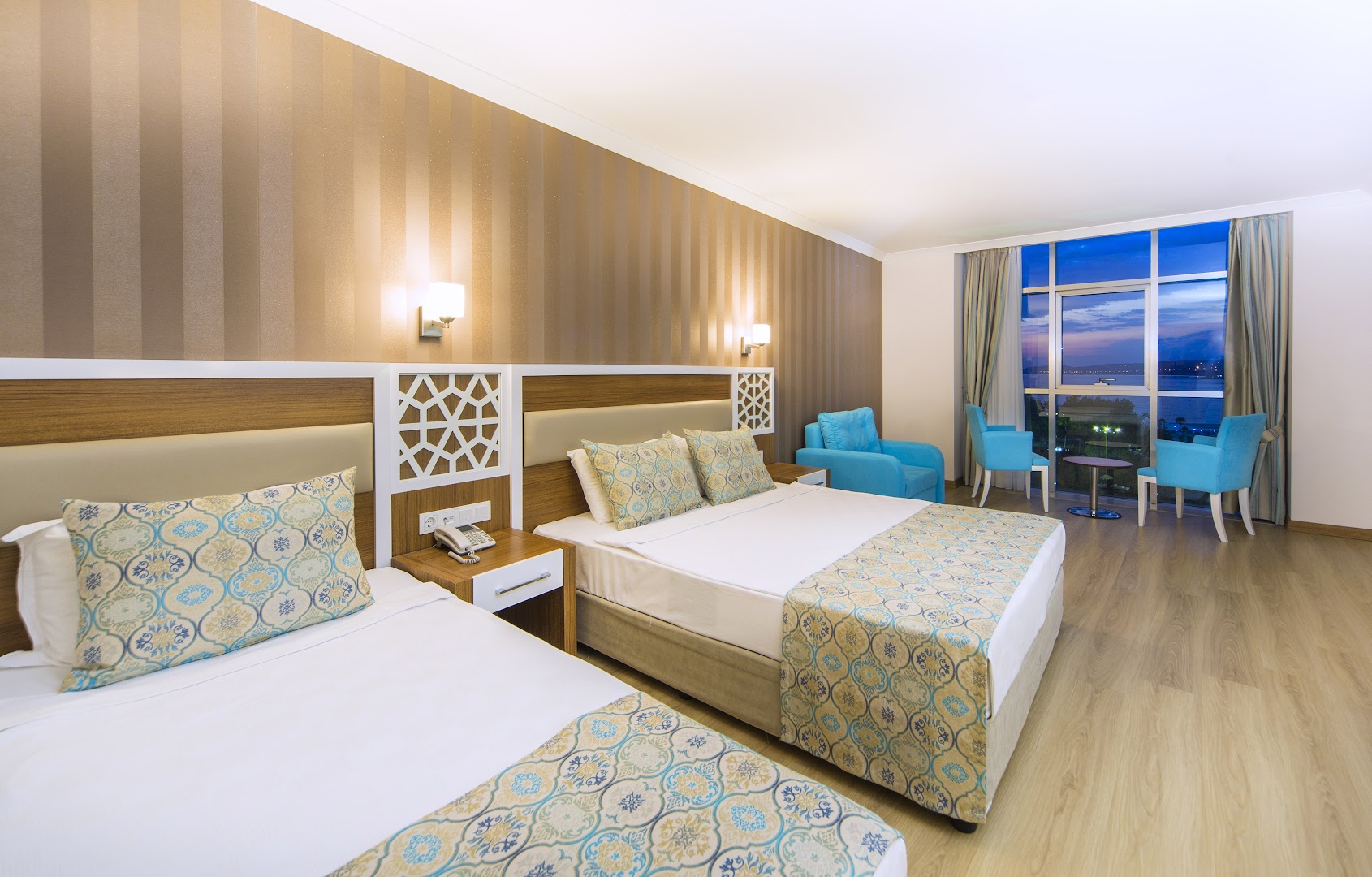 Lonicera Resort & Spa Hotel gaišs numurs ar divām gultām, tirkīza krēsliem un panorāmas logu uz jūru saulrieta laikā Alanjā