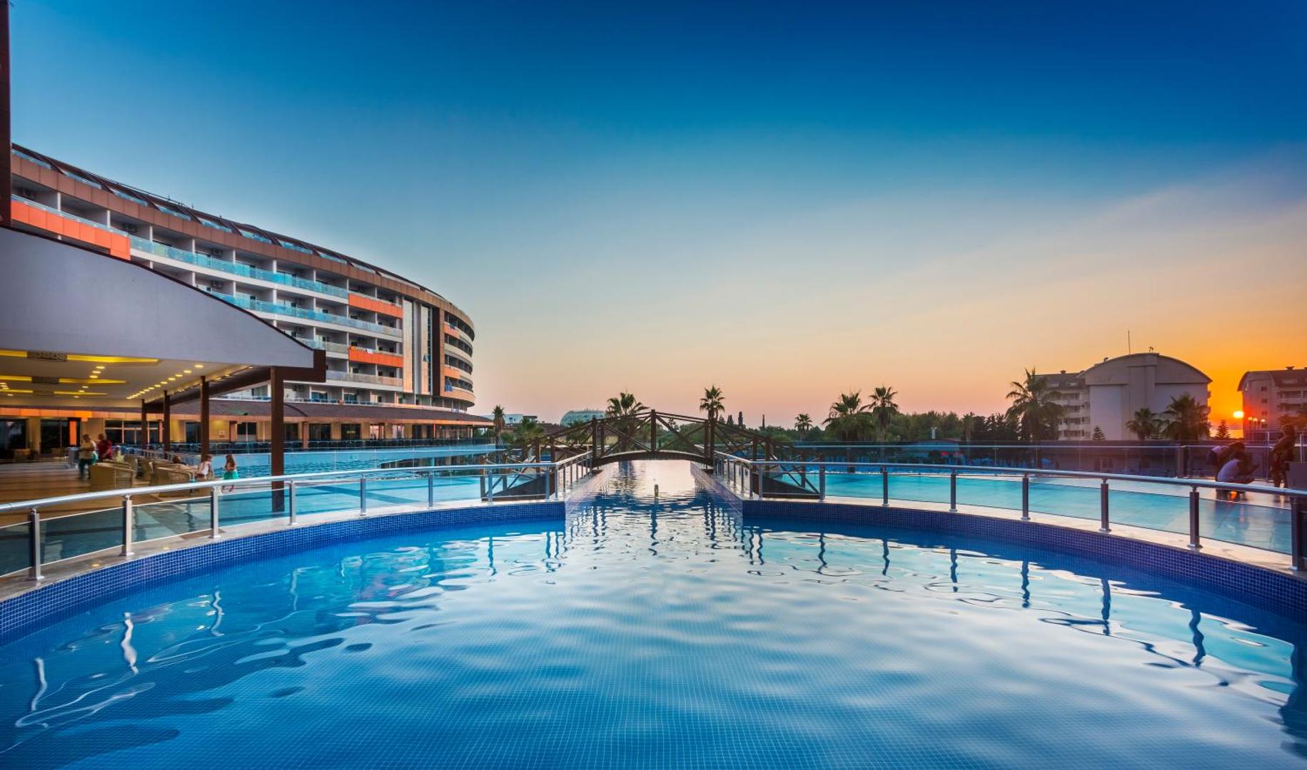 Lonicera Resort & Spa baseins saulrieta laikā ar dekoratīvu koka tiltiņu un skatu uz jūras horizontu Turcijā