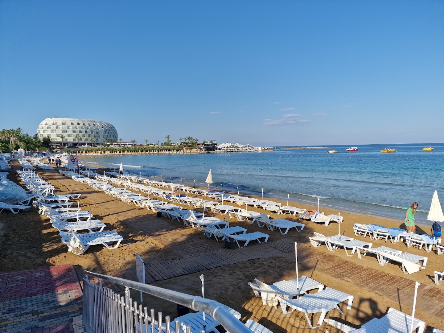 Lonicera Resort & Spa Hotel plašā smilšu pludmale Alanjā ar daudziem sauļošanās krēsliem, saulessargiem un skatu uz viesnīcas galveno ēku