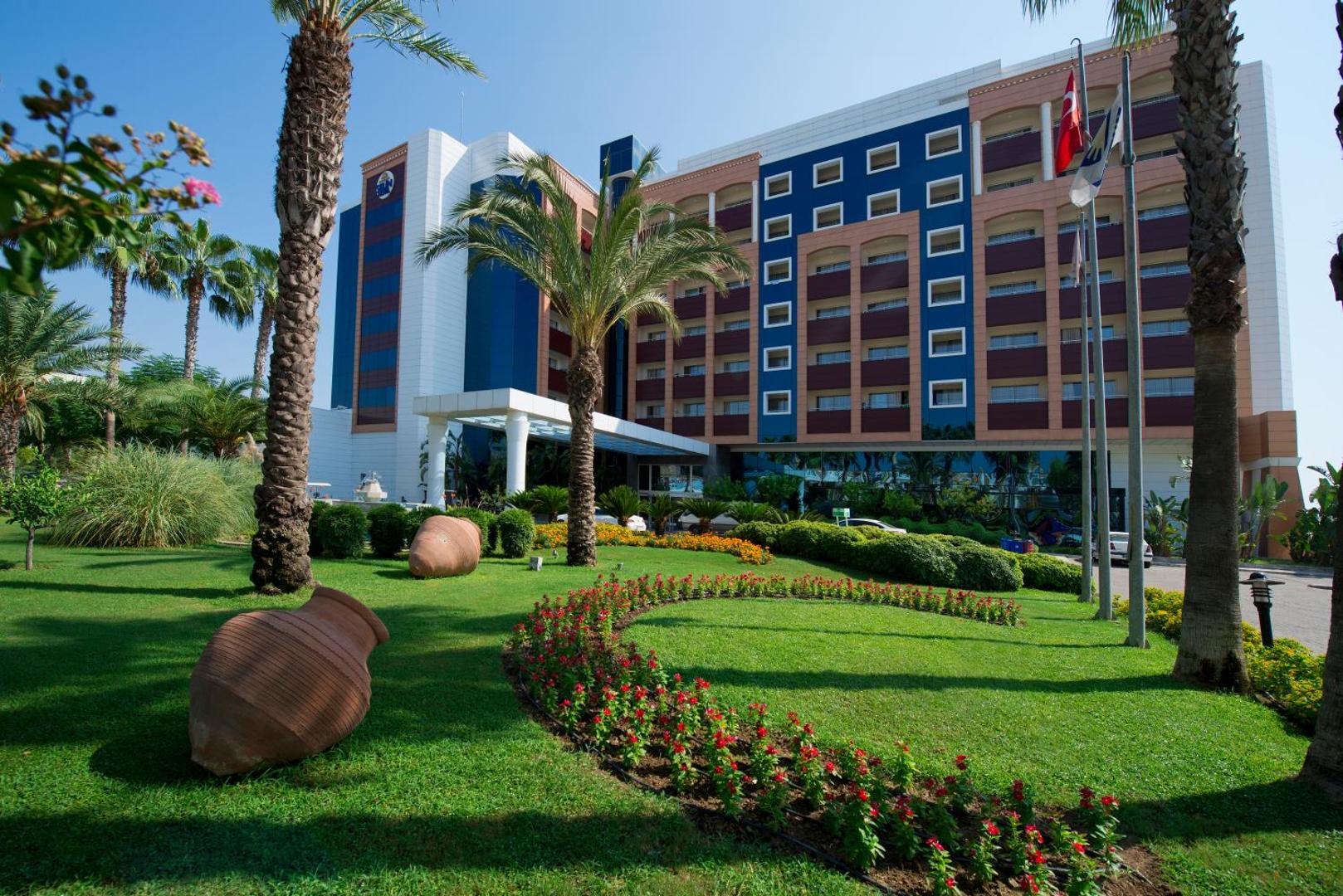Sentido Kamelya Selin Luxury Resort & SPA galvenā ēka un ieeja Sidē — sakopts dārzs ar palmām, ziediem un dekoratīvām amforām