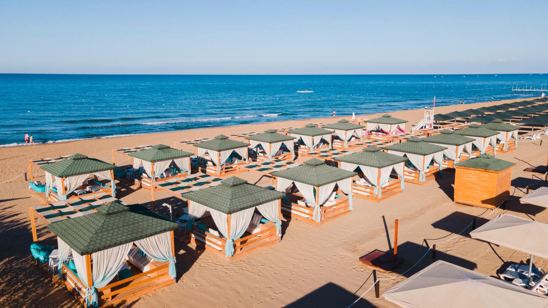 Sentido Kamelya Selin Luxury Resort & SPA plašā smilšu pludmale ar daudzām koka atpūtas nojumēm un zilo jūru Sidē