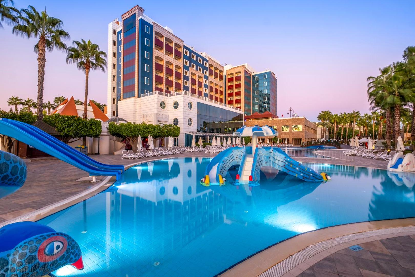 Sentido Kamelya Selin Luxury Resort & SPA bērnu baseins ar slidkalniņiem pie viesnīcas galvenās ēkas vakara apgaismojumā