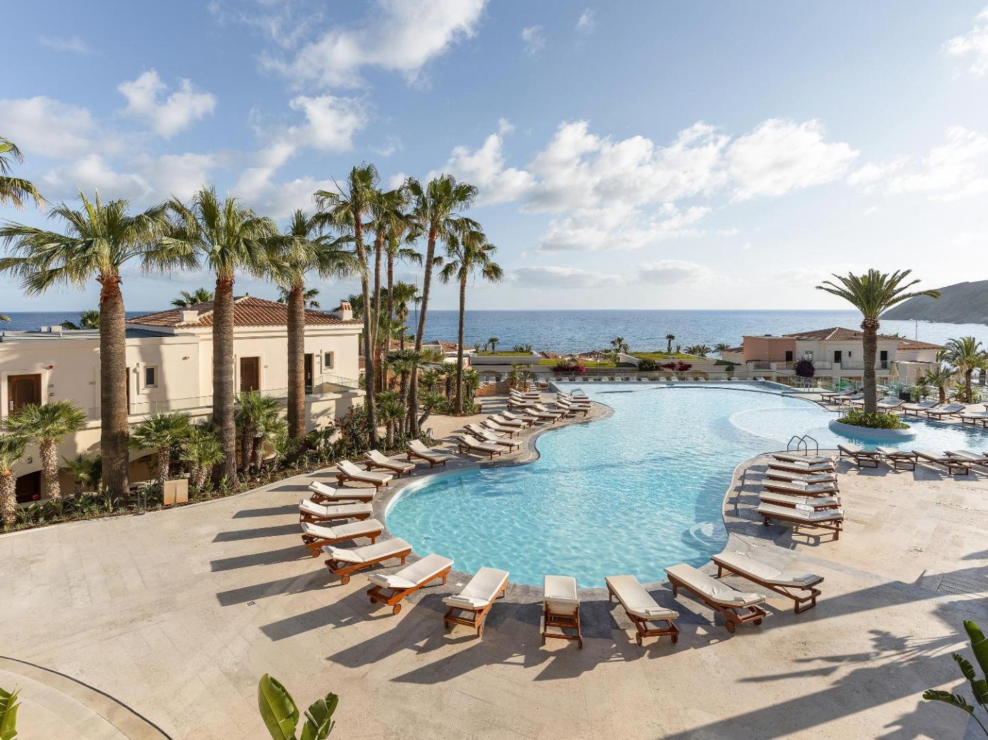 Grecotel Marine Palace & Aqua Park plašais centrālais baseins ar daudziem sauļošanās krēsliem un skatu uz jūru Panormo piekrastē