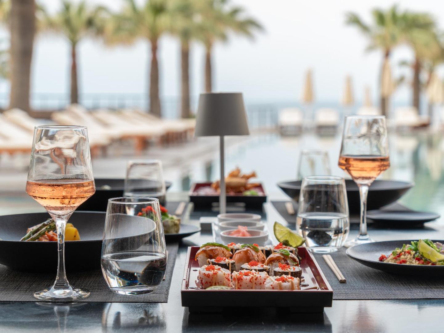 Grecotel Marine Palace & Aqua Park restorāna galds ar suši un vīnu pie āra baseina un palmām saulrietā