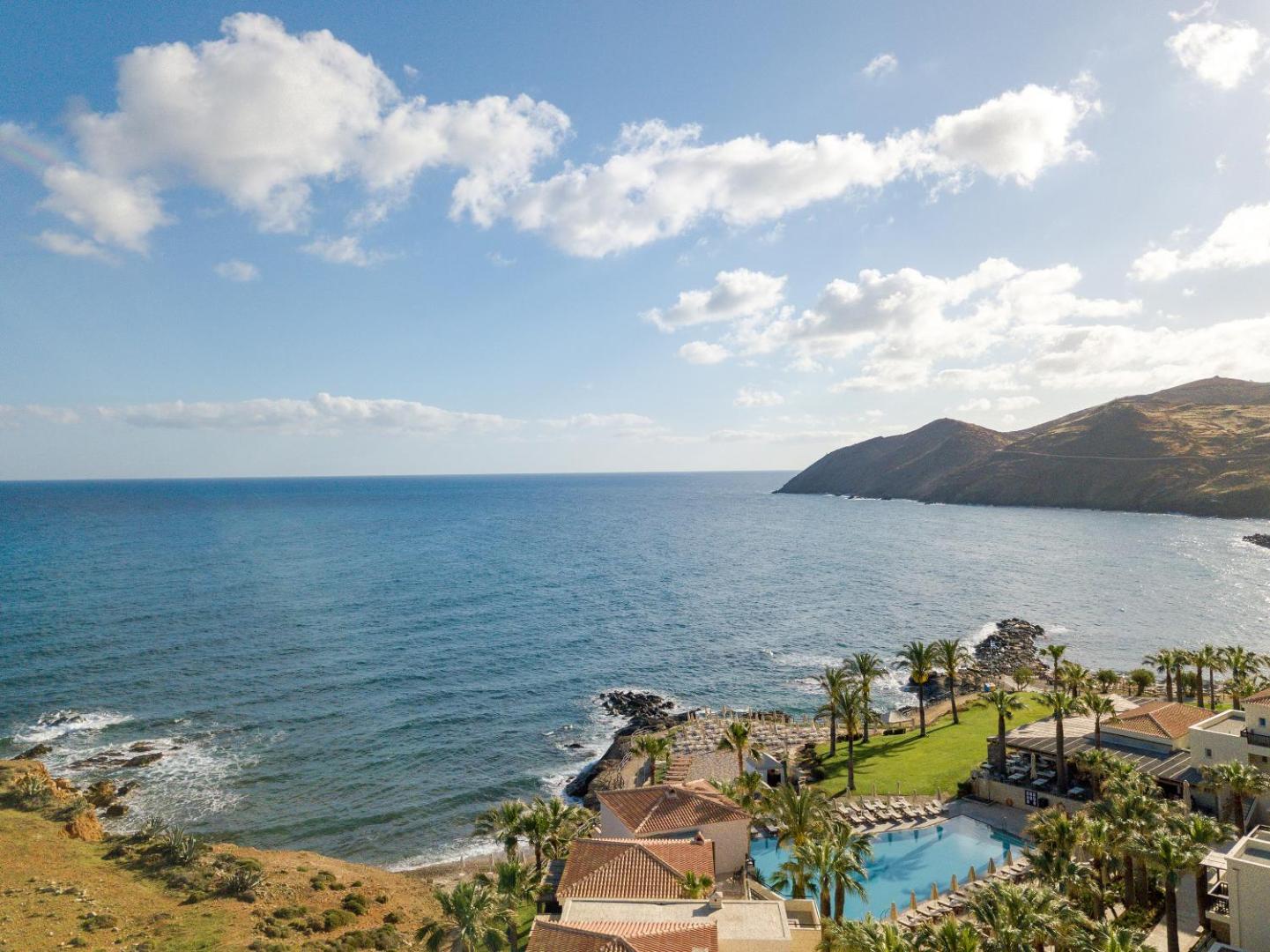 Grecotel Marine Palace & Aqua Park panorāmas skats uz piekrasti Krētā — āra baseins, palmas un viesnīcas ēkas pie jūras līča