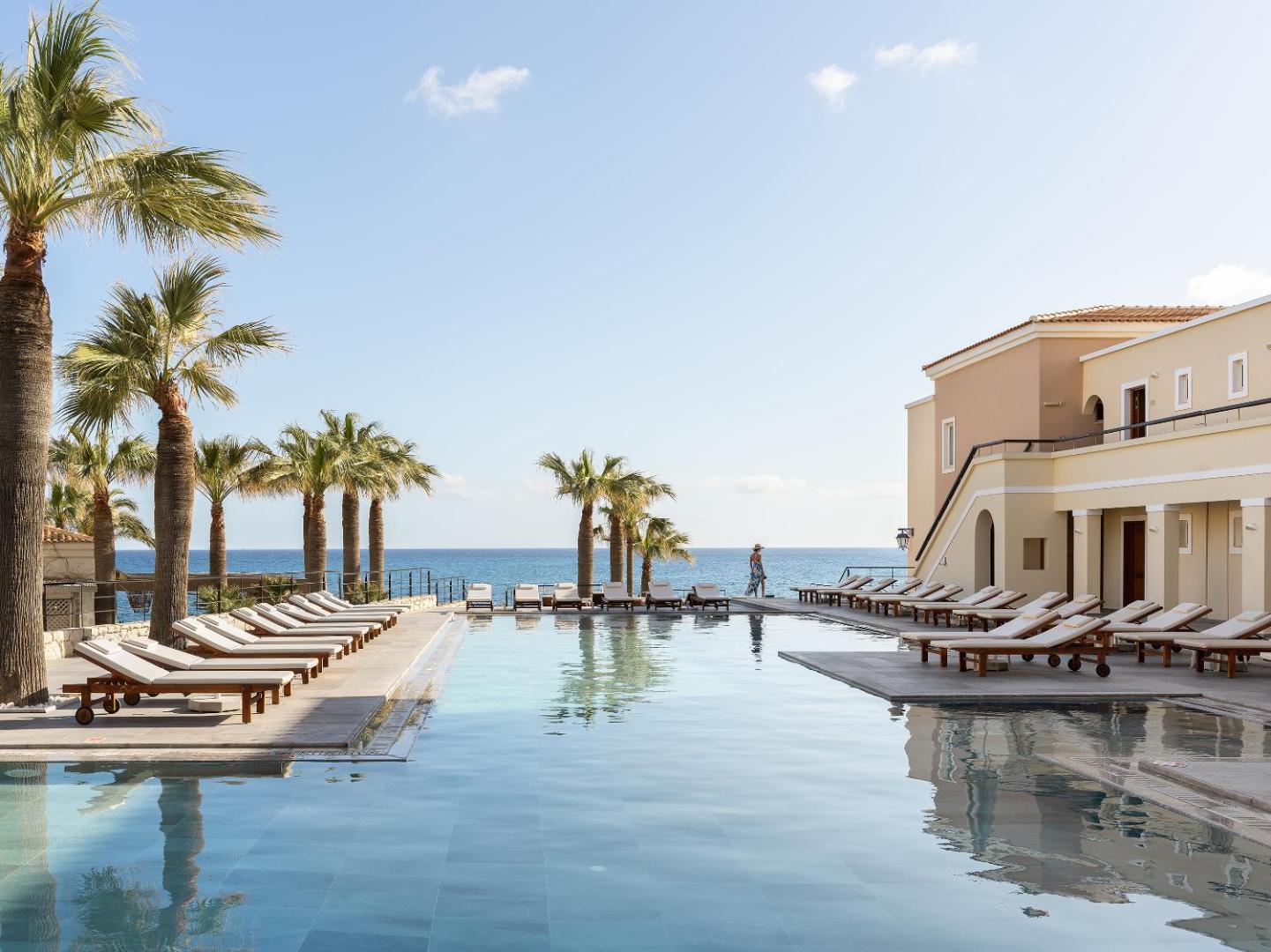 Grecotel Marine Palace & Aqua Park āra baseins ar skatu uz jūru Krētā — sauļošanās krēsli, palmas un viesnīcas ēka