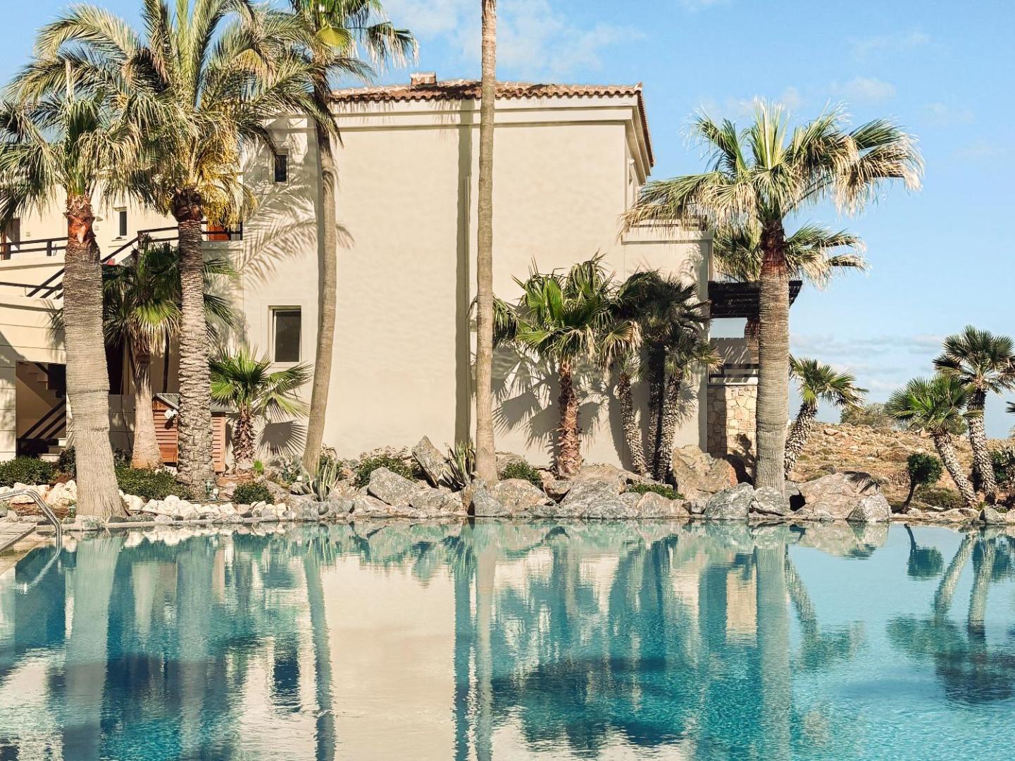 Grecotel Marine Palace & Aqua Park āra baseins ar palmu atspulgu ūdenī un viesnīcas korpusu fonā