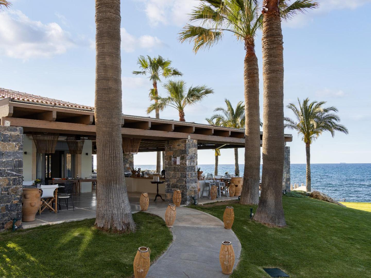 Grecotel Marine Palace & Aqua Park bāra terase ar akmens kolonnām un palmām — skats uz jūru un zālāju