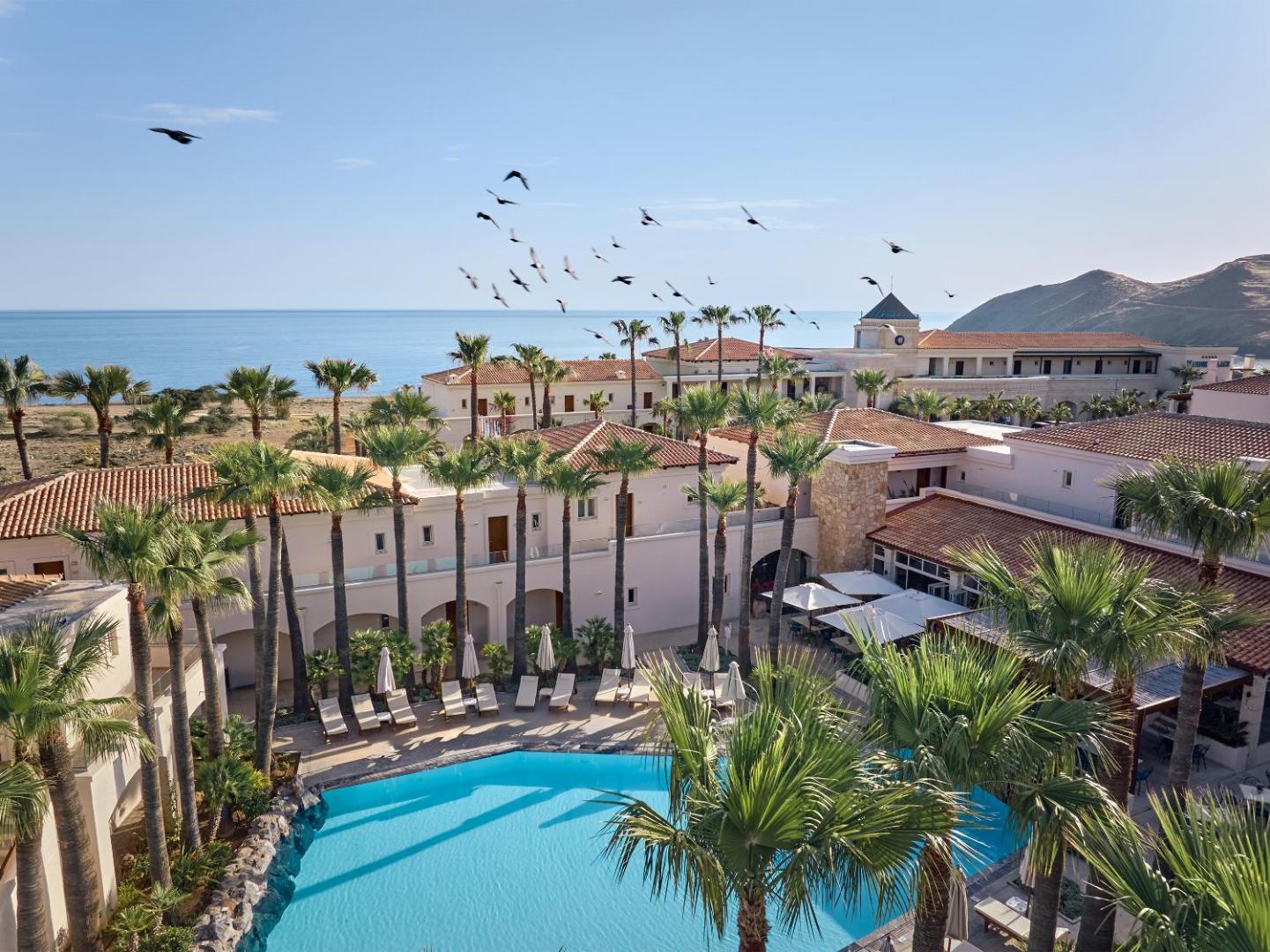 Grecotel Marine Palace & Aqua Park iekšpagalms ar baseinu, palmām un atpūtas krēsliem piekrastē zem skaidrām debesīm