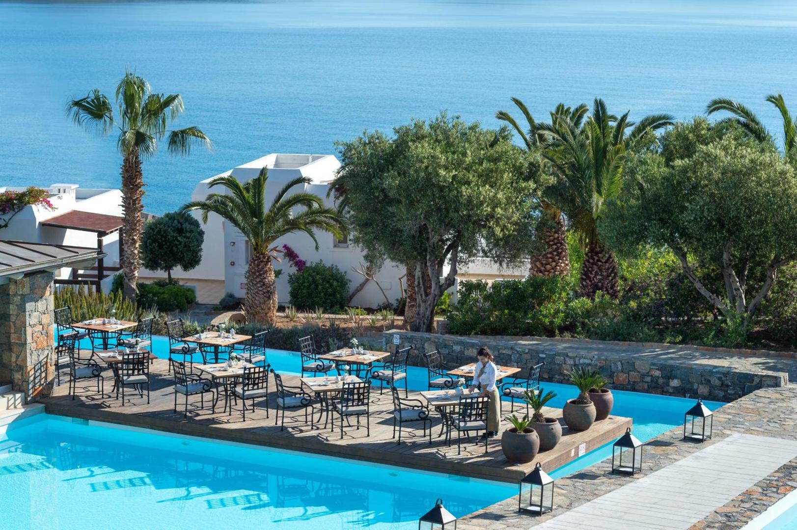 Aquila Elounda Village āra restorāna terase uz koka klāja virs zila baseina, fonā Vidusjūra un palmas Krētā