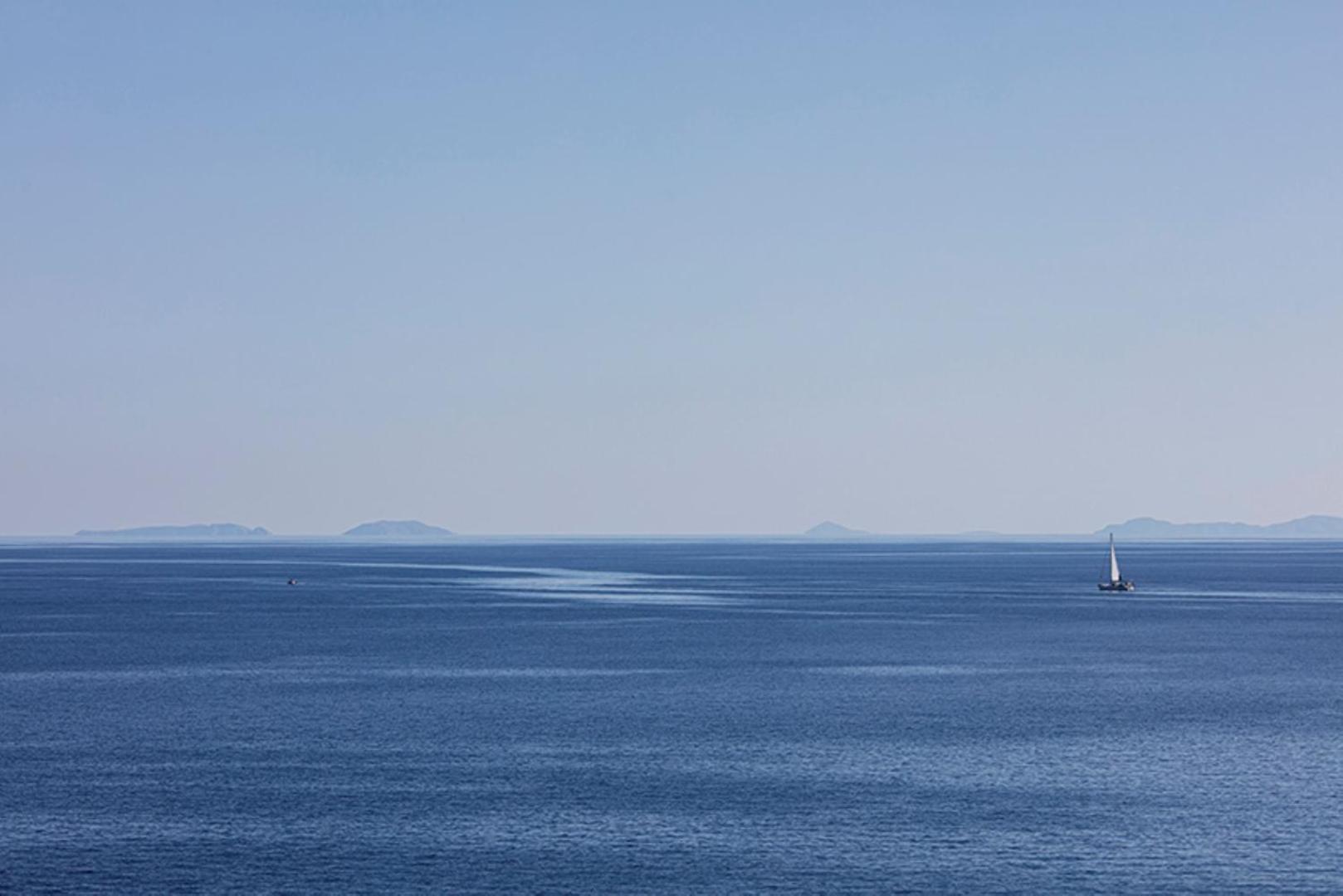 Skats uz zilo jūru un tālumā esošajām salām no Aquila Elounda Village — neliela jahta burā pa mierīgu ūdeni