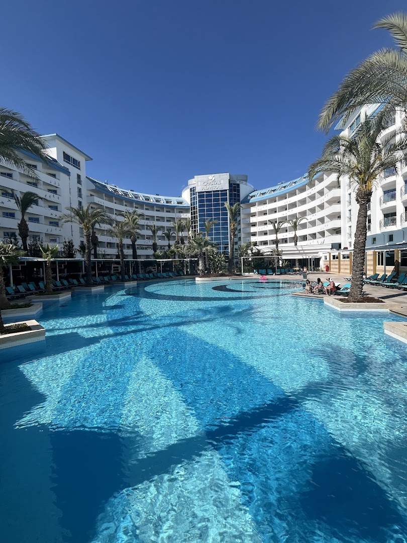Crystal Admiral Resort Suites & Spa modernā arhitektūra un lielais baseins ar palmām saulainā dienā Turcijas kūrortā