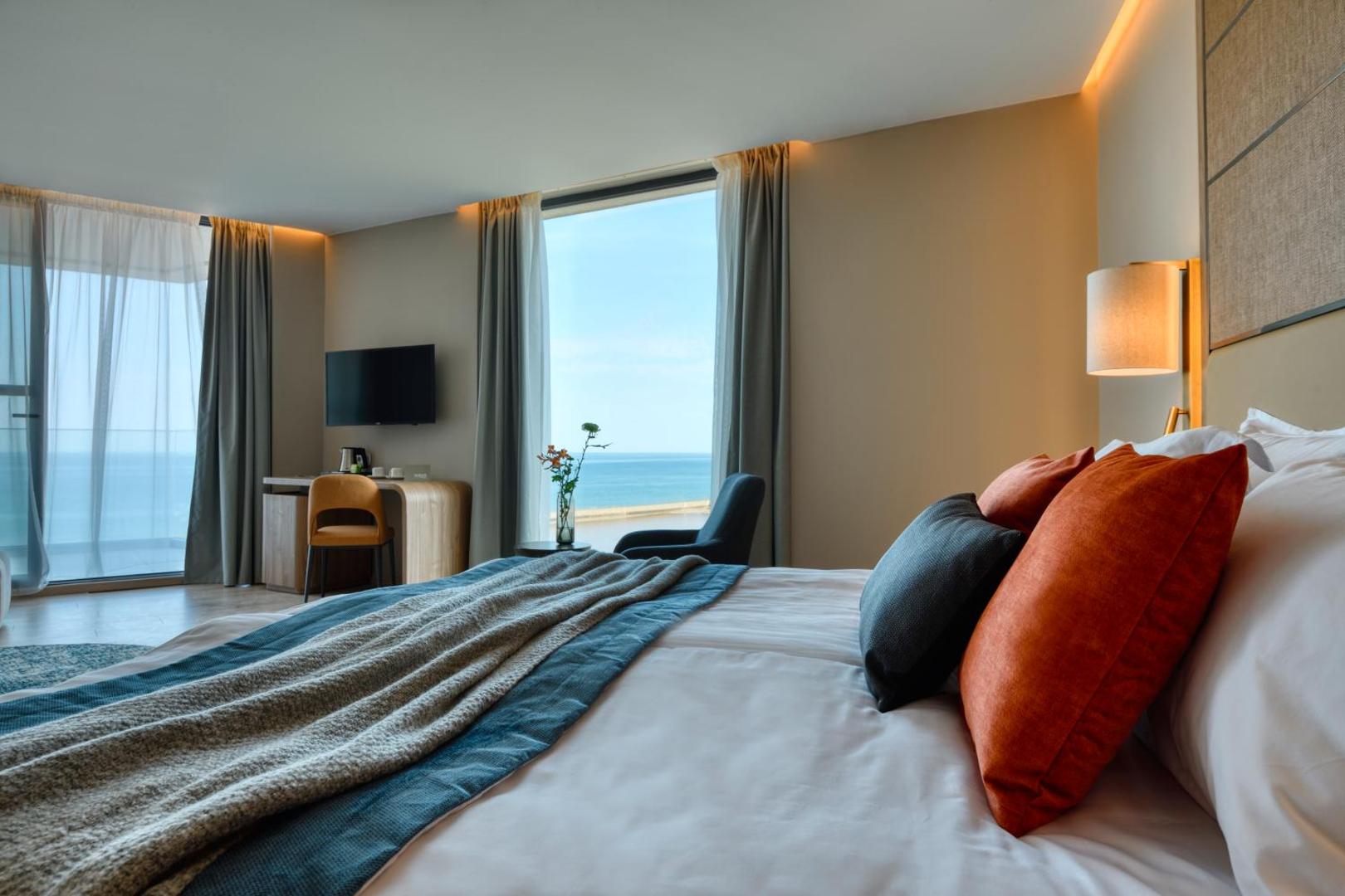 The Wave Resort numura interjers ar skatu uz Melno jūru caur logu, fokusā gulta ar dekoratīviem spilveniem