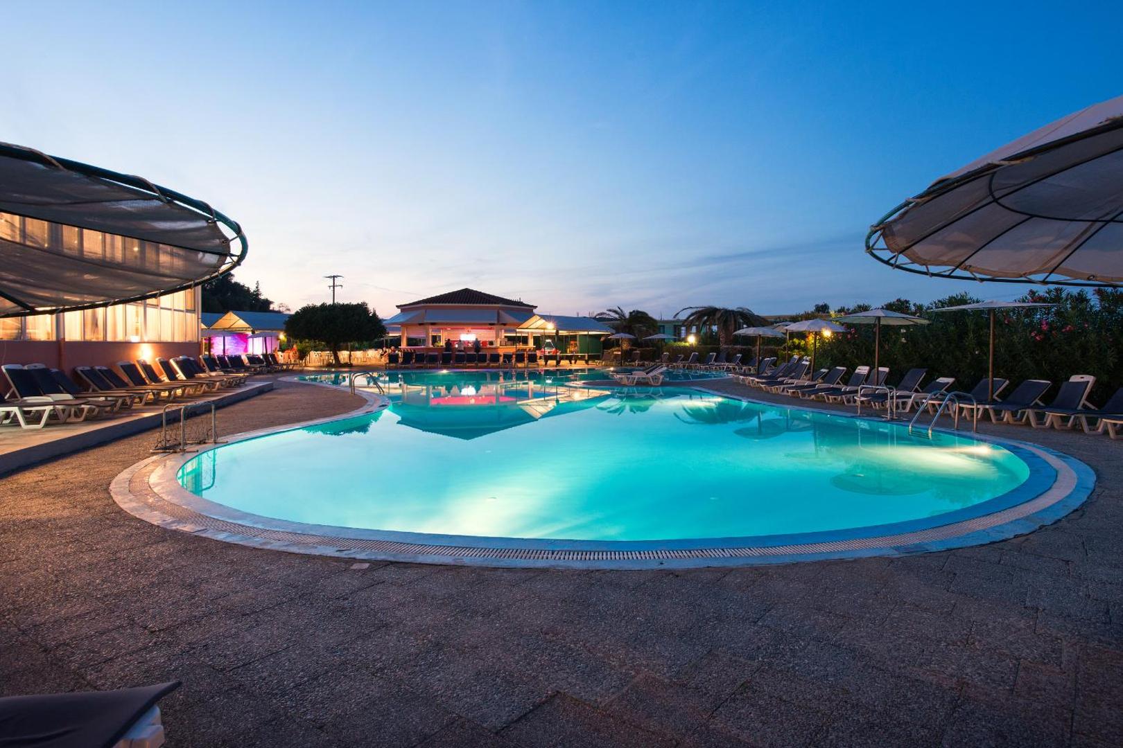 Panorama Sidari Hotel āra baseins krēslas stundā ar izgaismotu ūdeni un sauļošanās krēsliem Korfu salas ziemeļos