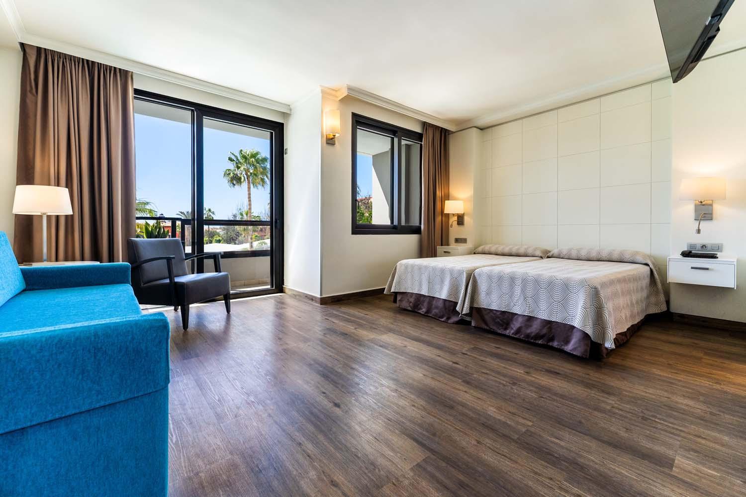 Gaišs Hotel Bitacora viesnīcas numurs ar divām gultām, zilu dīvānu, koka grīdu un balkonu ar skatu uz palmām.
