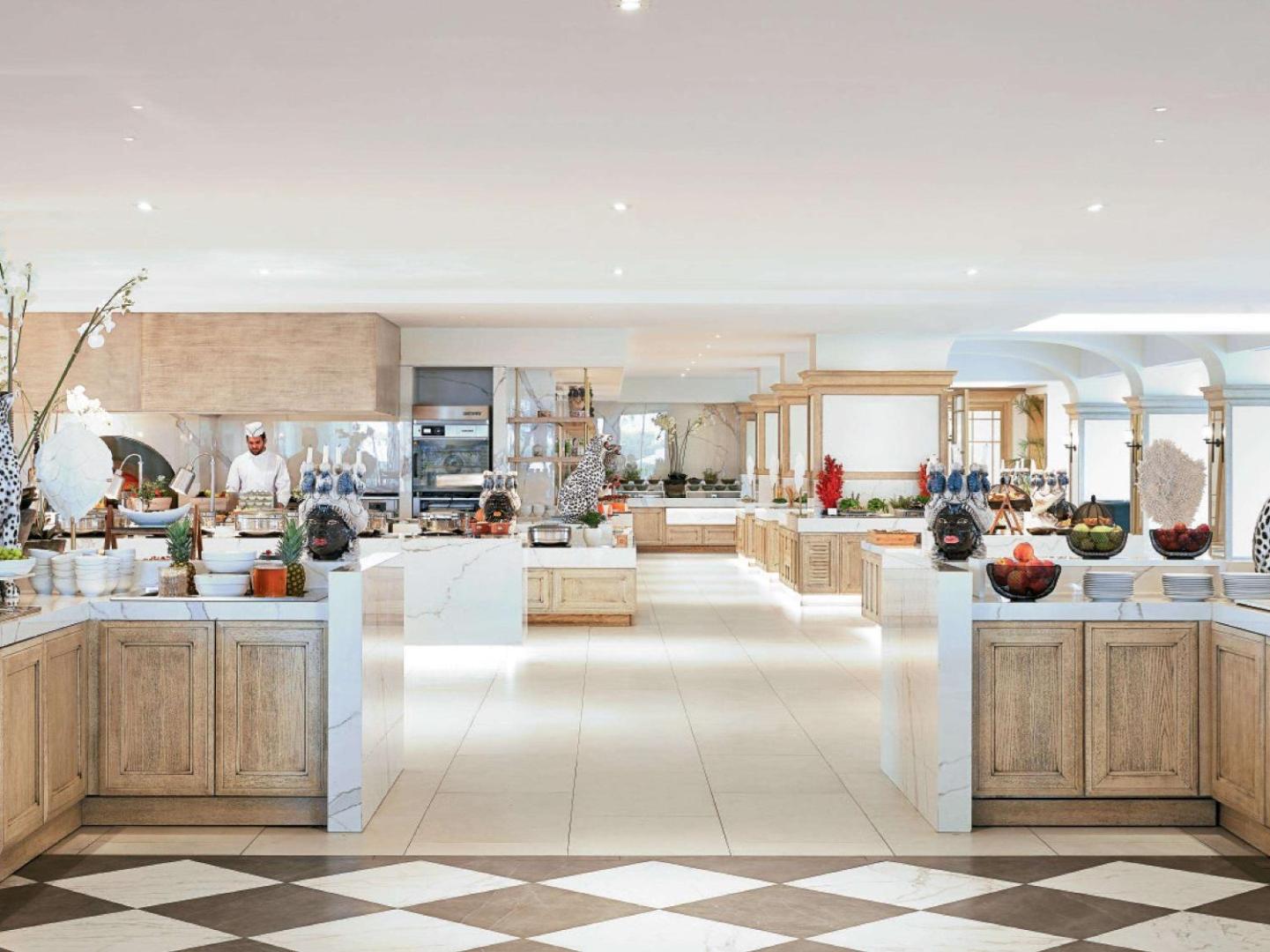 Corfu Imperial Grecotel Exclusive Resort galvenais restorāns ar plašu bufetes zonu, gaišu interjeru un šefpavāru pie atvērtās virtuves stacijas.