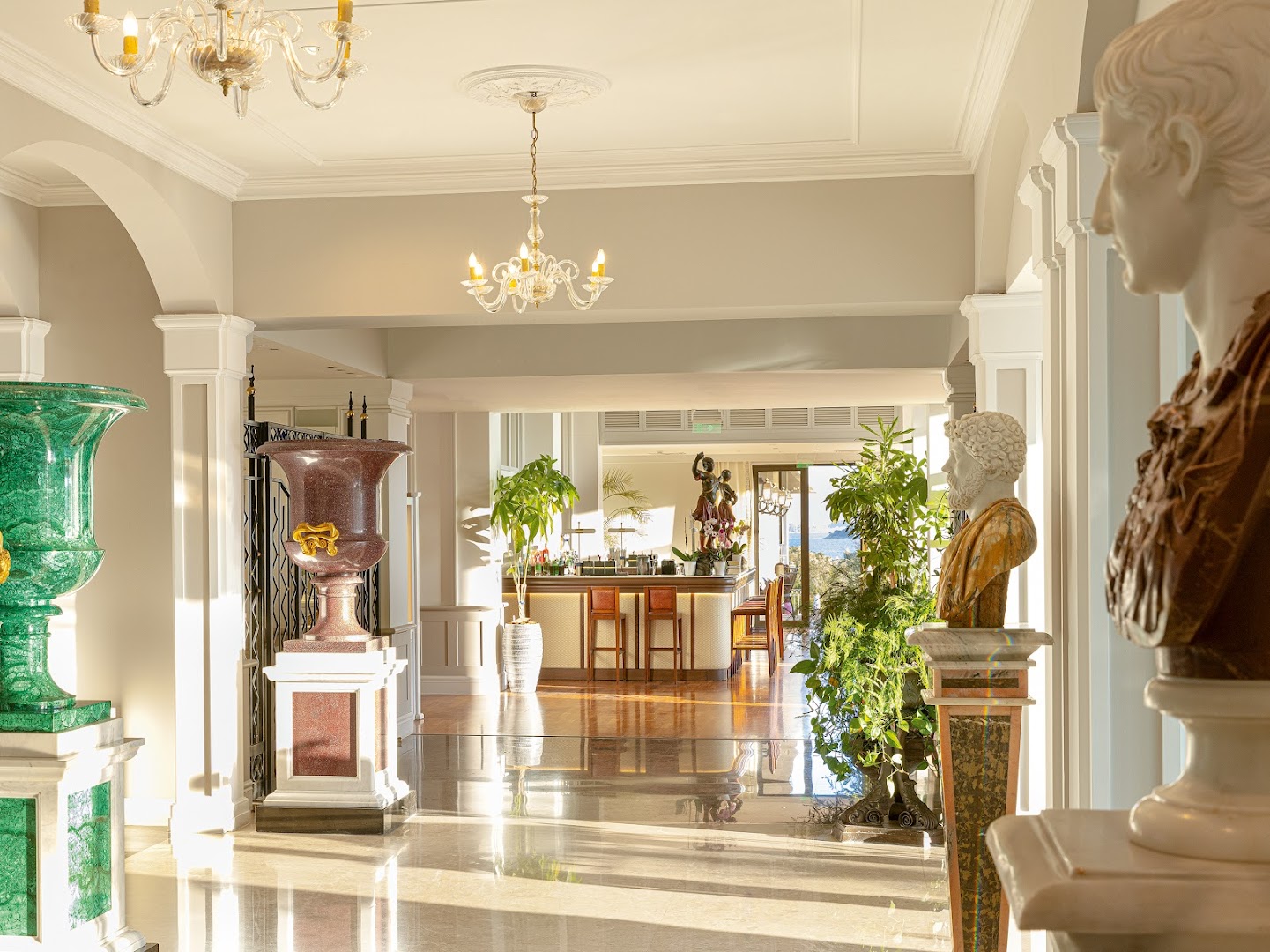 Corfu Imperial Grecotel viesnīcas vestibils ar marmora grīdām, antīkām skulptūrām un skatu uz bāru un jūru fonā.