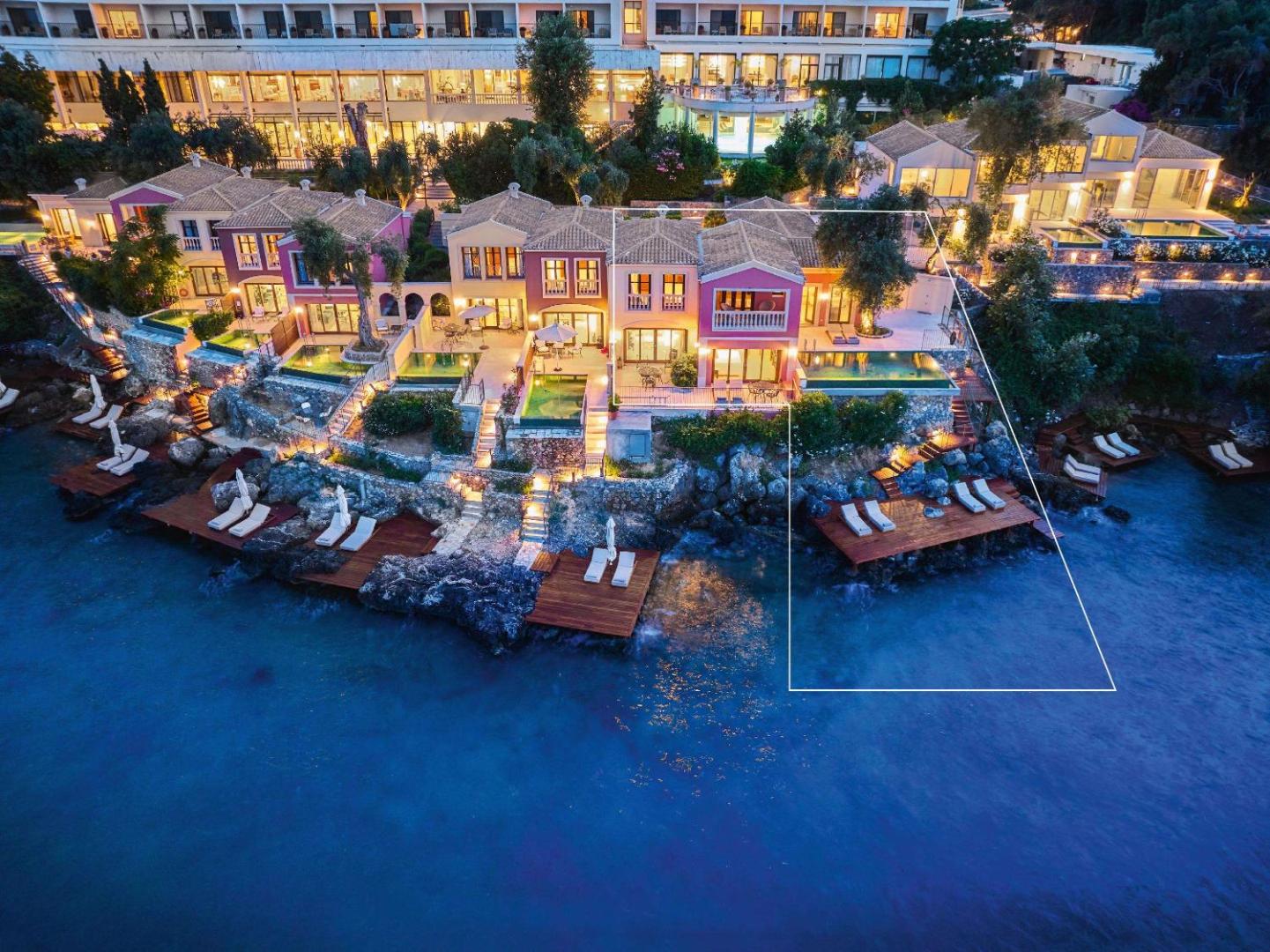 Corfu Imperial Grecotel Exclusive Resort piekrastes villas ar privātiem baseiniem un koka terasēm vakara apgaismojumā pie pašas jūras.