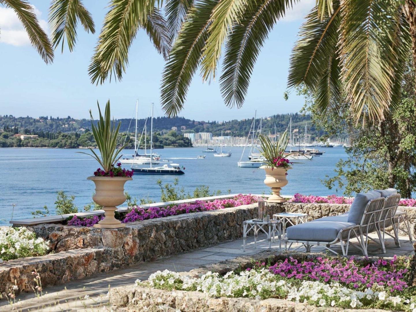 Corfu Imperial Grecotel Exclusive Resort terase ar skatu uz Jonijas jūru un jahtām — atpūtas zona ar dīvāniem un ziediem