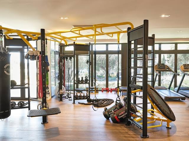 Corfu Imperial Grecotel mūsdienīgs fitness centrs ar trenažieriem, boksa maisu un funkcionālā treniņa aprīkojumu gaišā zālē ar skatu uz dārzu