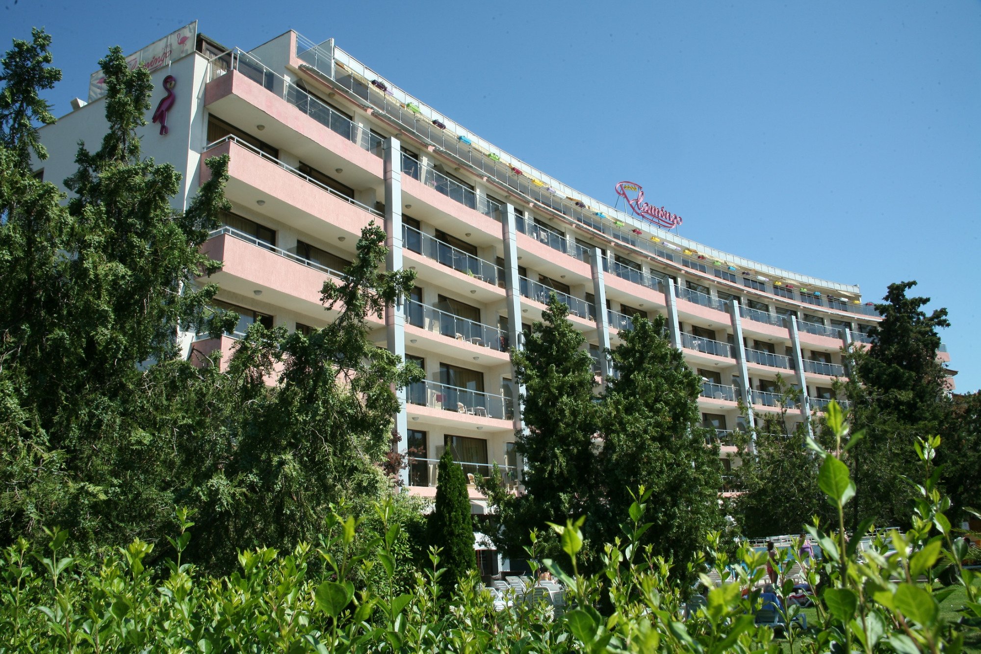 Flamingo Beach Hotel daudzstāvu ēkas fasāde ar balkoniem un rozā akcentiem, ko ieskauj zaļi koki un dārzs