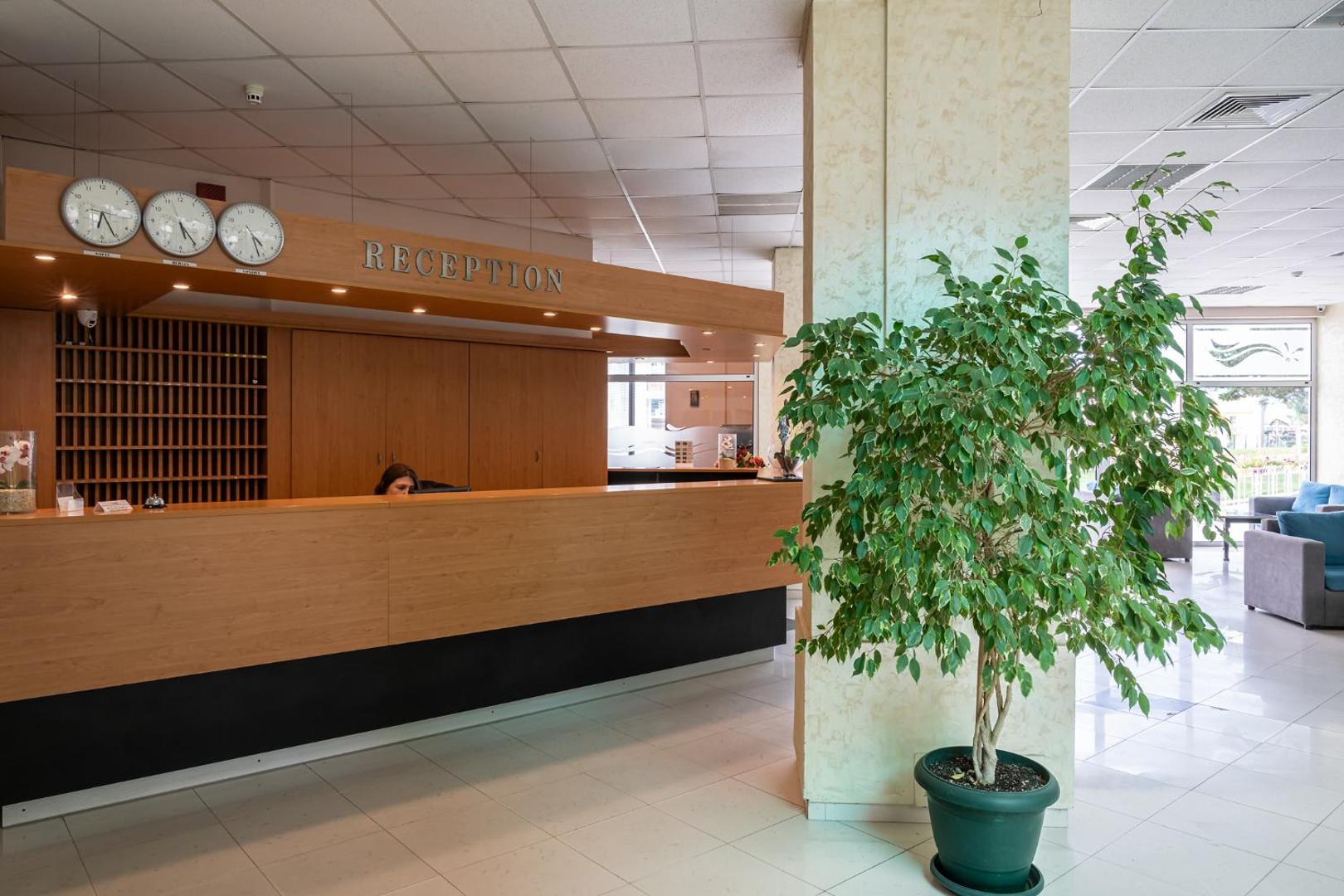 Flamingo Beach Hotel recepcijas zona ar koka apdari, pasaules pulksteņiem un telpaugu Larnakā, Kiprā — gaišs un plašs vestibils