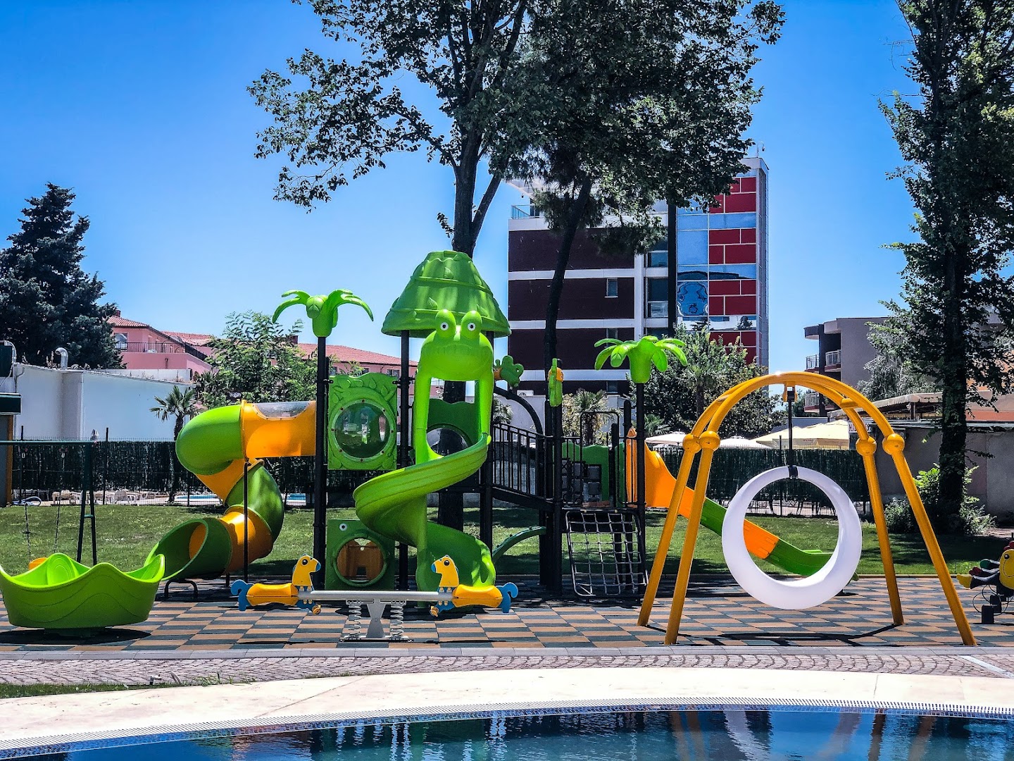 Flamingo Beach Hotel bērnu rotaļu laukums ar krāsainiem slidkalniņiem un šūpolēm pie baseina — droša izklaides zona ģimenēm ar bērniem