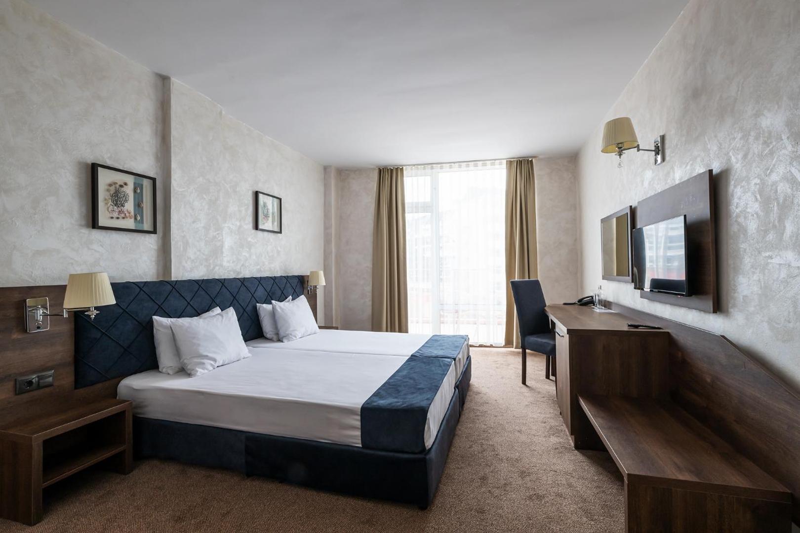 Flamingo Beach Hotel gaišs divvietīgs numurs ar tumši zilu gultas galvgalī, rakstāmgaldu, televizoru un lielu logu